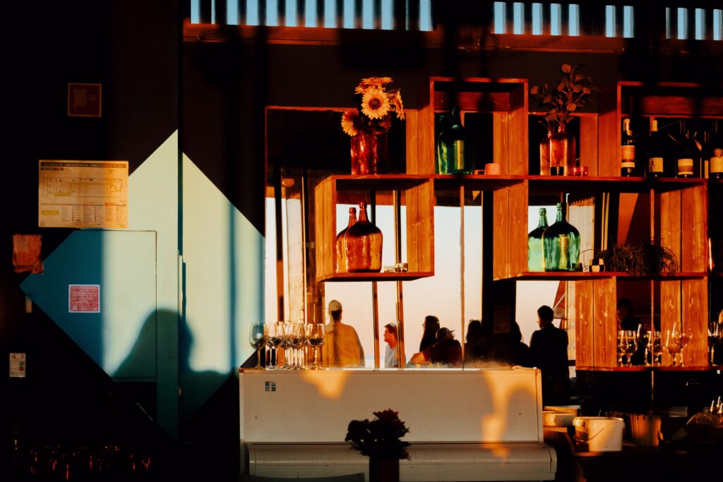A warm, sunlit bar in Lisboa with colorful bottles, a piano, and people silhouettes.